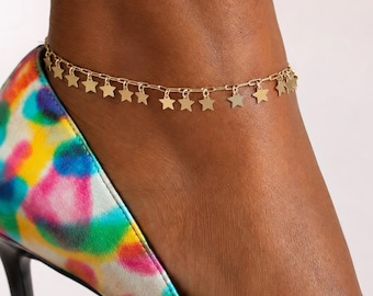 May include: A gold-toned anklet featuring a delicate chain adorned with a series of small, evenly spaced star-shaped charms. The anklet is worn around the ankle, positioned above a high-heeled pump with a multi-colored, abstract paint-splatter pattern.