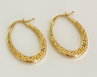 以下が含まれることがあります： 金色の楕円形フープピアス2つ。各イヤリングは外縁にギリシャキーのデザインが施されています。イヤリングは白い背景に表示されています。
