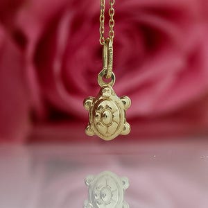 Op de afbeelding: Een gouden schildpadhanger aan een fijne ketting. De schildpadbedel heeft een gedetailleerd schildpatroon en kleine pootjes. De achtergrond is wazig roze en wit.