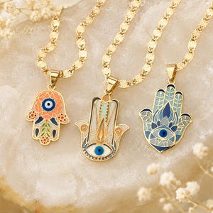 May include: Three gold-tone necklaces featuring hamsa hand pendants with colorful enamel detailing. Each pendant includes a central blue and white evil eye motif. The chains consist of flat, textured gold-tone links with a reflective finish.