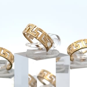 May include: Three gold rings with a Greek key design on a clear display stand. The rings are stacked on top of each other.
