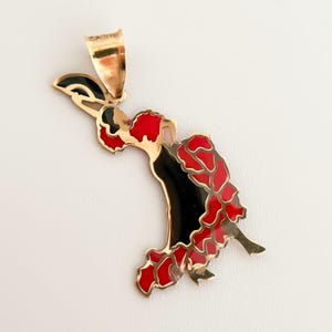 May include: Gold-tone pendant featuring a flamenco dancer design. The dancer's dress is black and red, with intricate detailing. The pendant has a bail for attaching to a chain. The overall design is elegant and detailed.