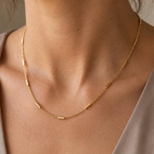 Puede incluir: Un collar de cadena dorada que presenta peque&ntilde;os acentos de barras met&aacute;licas cil&iacute;ndricas espaciadas uniformemente a lo largo de la delicada cadena de eslabones. El collar descansa sobre el cuello, mostrando un dise&ntilde;o simple y pulido.