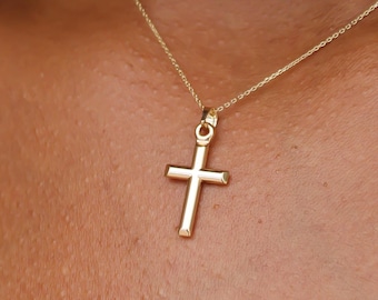May include: A gold-toned metal cross pendant suspended from a delicate, thin gold-toned chain. The cross features a smooth, polished surface with a simple, clean design. The necklace rests against the neck, showcasing the metallic luster of the gold.