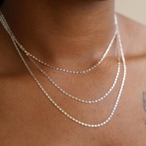 Puede incluir: Tres delicados collares plateados de diferentes longitudes se superponen en el pecho de una persona. Los collares tienen un dise&ntilde;o simple y elegante, con peque&ntilde;os eslabones planos. La persona lleva una camiseta negra y un tatuaje de mariposa es visible en su hombro.