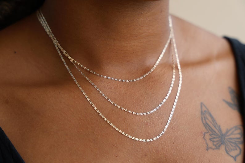 Puede incluir: Tres delicados collares plateados de diferentes longitudes se superponen en el pecho de una persona. Los collares tienen un dise&ntilde;o simple y elegante, con peque&ntilde;os eslabones planos. La persona lleva una camiseta negra y un tatuaje de mariposa es visible en su hombro.