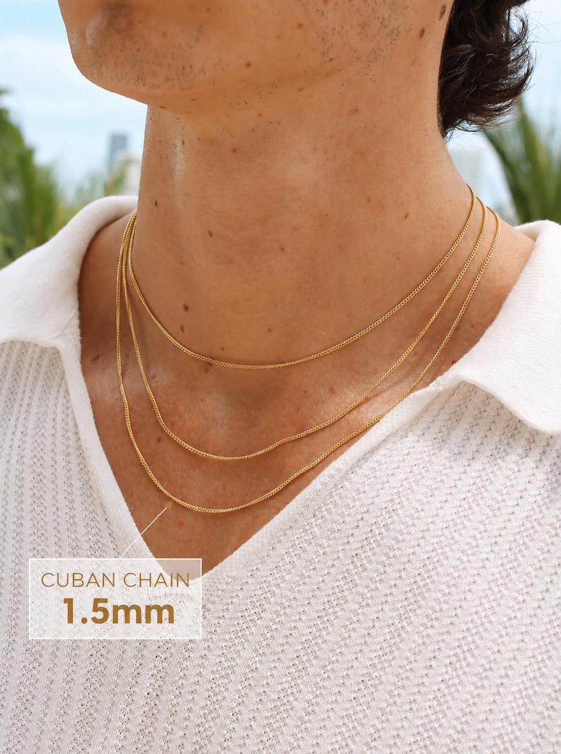 May include: Three layered gold-toned Cuban chains worn around the neck. The chains feature a delicate link design with a thickness of 1.5mm as indicated by the text overlay. The necklaces are worn over a white textured collared shirt.