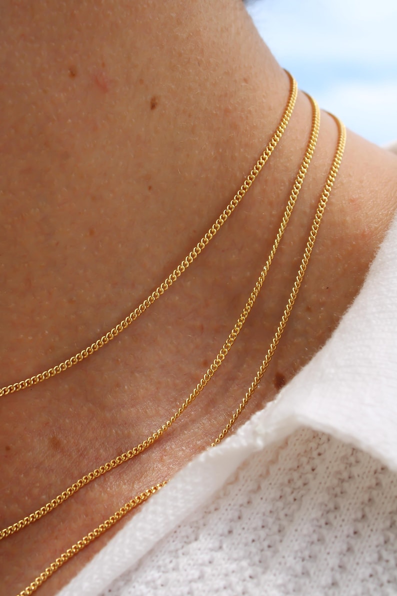 May include: A set of three delicate gold chain necklaces worn layered around the neck. Each necklace features a fine, uniform link design with a polished, reflective finish that catches the light, creating a subtle and elegant layered appearance.
