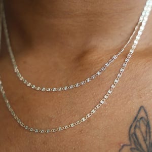Puede incluir: Primer plano de dos delicados collares de plata con un dise&ntilde;o de eslabones de cadena. Los collares est&aacute;n superpuestos, uno ligeramente m&aacute;s largo que el otro. Las cadenas se muestran sobre un fondo de tono de piel.