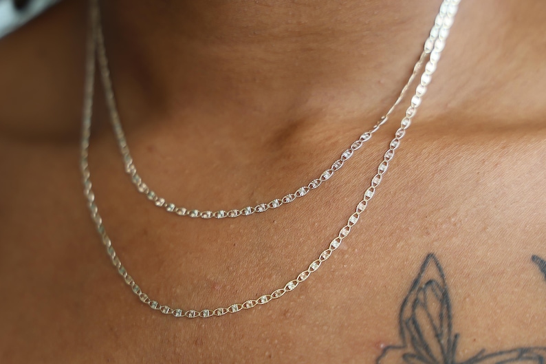 Puede incluir: Primer plano de dos delicados collares de plata con un dise&ntilde;o de eslabones de cadena. Los collares est&aacute;n superpuestos, uno ligeramente m&aacute;s largo que el otro. Las cadenas se muestran sobre un fondo de tono de piel.