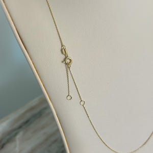 May include: A delicate gold necklace with a fine chain and adjustable length. The necklace features a small clasp and two adjustment rings. The gold chain is displayed on a cream-colored surface.
