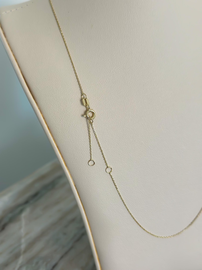May include: A delicate gold necklace with a fine chain and adjustable length. The necklace features a small clasp and two adjustment rings. The gold chain is displayed on a cream-colored surface.