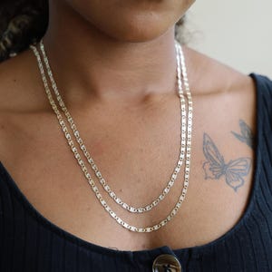Puede incluir: Se muestran dos collares plateados con un dise&ntilde;o de eslabones rectangulares. Los collares est&aacute;n superpuestos, uno ligeramente m&aacute;s largo que el otro. Los collares se llevan sobre una camiseta sin mangas negra.