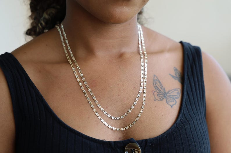 Puede incluir: Se muestran dos collares plateados con un dise&ntilde;o de eslabones rectangulares. Los collares est&aacute;n superpuestos, uno ligeramente m&aacute;s largo que el otro. Los collares se llevan sobre una camiseta sin mangas negra.