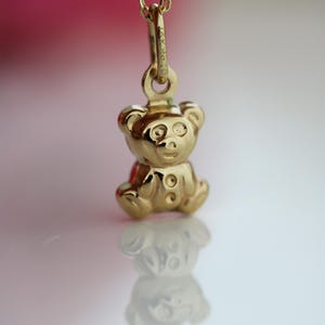 Op de afbeelding: Een gouden teddybeer hanger aan een fijne ketting. De hanger, een miniatuur beer met knoopdetails, is zeer reflecterend. De achtergrond is zacht, met de reflectie van de hanger eronder zichtbaar.