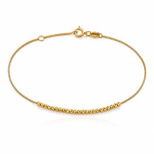 May include: A gold chain bracelet featuring a delicate box chain design with a row of small, round, textured gold beads centered along the strand. The bracelet is secured with a circular spring ring clasp and includes a small jump ring for adjustment.