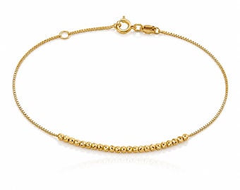 May include: A gold chain bracelet featuring a delicate box chain design with a row of small, round, textured gold beads centered along the strand. The bracelet is secured with a circular spring ring clasp and includes a small jump ring for adjustment.