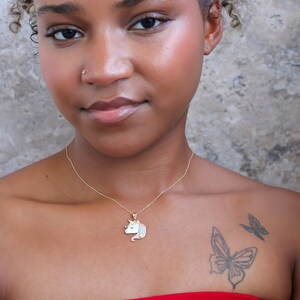 May include: A gold-colored necklace with a unicorn pendant. The pendant features a white, pink, and blue unicorn design. The person in the image is wearing a red top and has a butterfly tattoo on their chest.