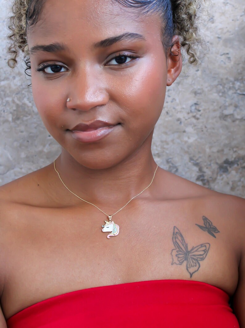 May include: A gold-colored necklace with a unicorn pendant. The pendant features a white, pink, and blue unicorn design. The person in the image is wearing a red top and has a butterfly tattoo on their chest.