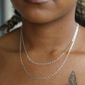 Puede incluir: Dos delicados collares de cadena de plata superpuestos en el cuello de una persona. Las cadenas tienen un dise&ntilde;o simple y elegante. Un tatuaje de mariposa es visible en el pecho de la persona. El fondo est&aacute; borroso.