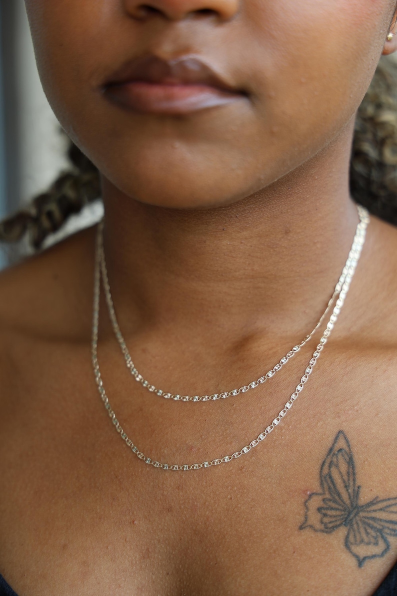 Puede incluir: Dos delicados collares de cadena de plata superpuestos en el cuello de una persona. Las cadenas tienen un dise&ntilde;o simple y elegante. Un tatuaje de mariposa es visible en el pecho de la persona. El fondo est&aacute; borroso.