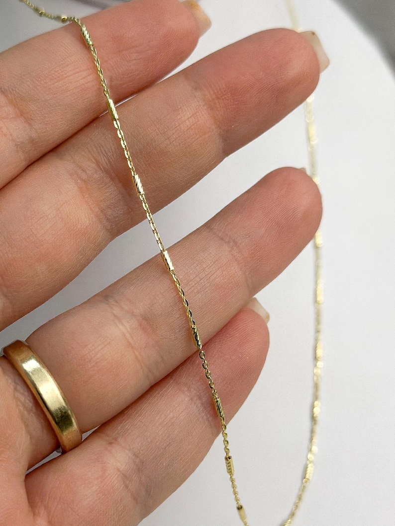 Puede incluir: Un delicado collar de cadena de oro que presenta una serie de peque&ntilde;os eslabones cil&iacute;ndricos texturizados intercalados a lo largo de una fina cadena de eslabones pulidos. El metal tiene un acabado en oro amarillo brillante.