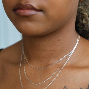 Puede incluir: Primer plano de tres delicados collares plateados superpuestos en el cuello de una persona. Los collares tienen diferentes estilos de cadena, incluyendo una cadena plana y rectangular. El tono de piel es marr&oacute;n medio, y un tatuaje de mariposa es visible.
