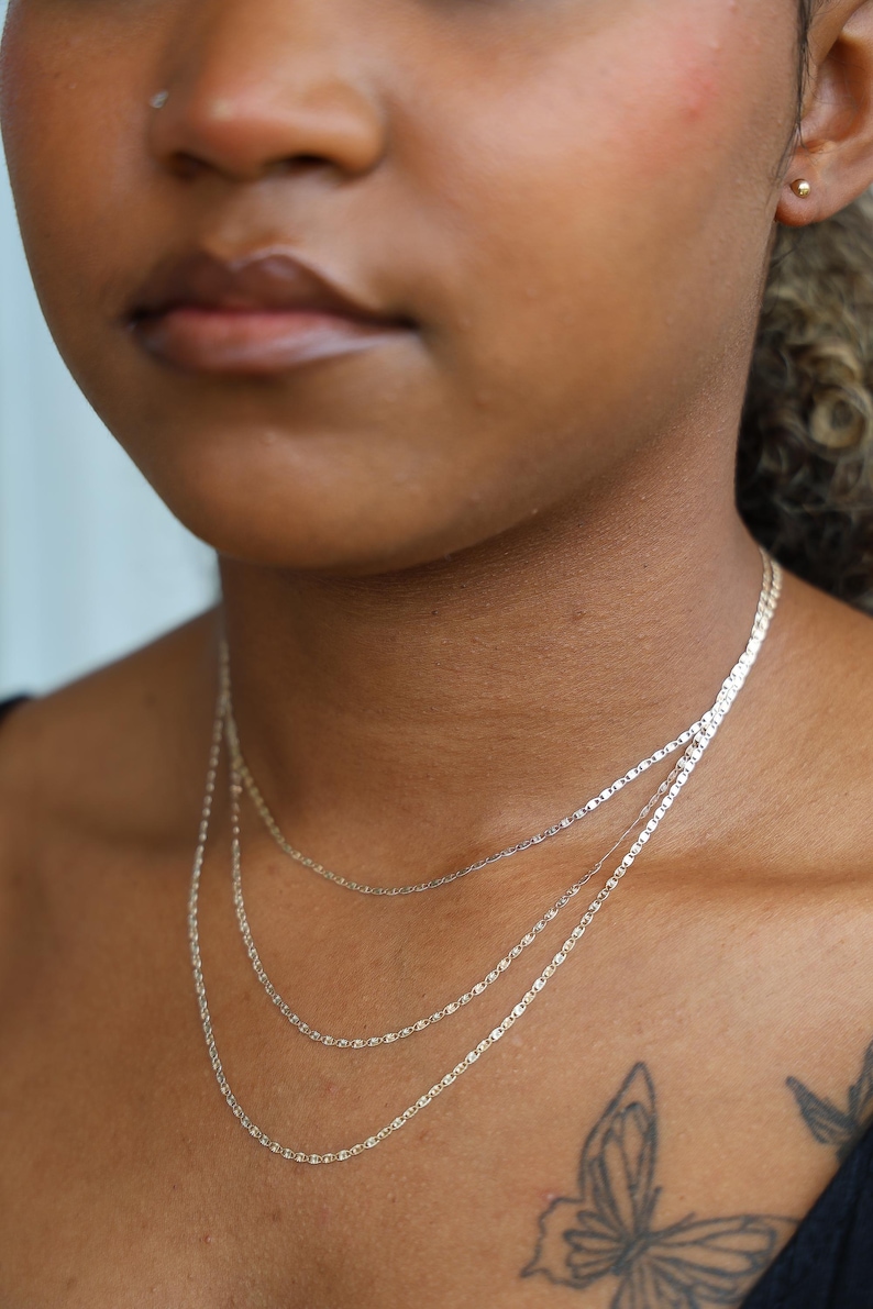 Puede incluir: Primer plano de tres delicados collares plateados superpuestos en el cuello de una persona. Los collares tienen diferentes estilos de cadena, incluyendo una cadena plana y rectangular. El tono de piel es marr&oacute;n medio, y un tatuaje de mariposa es visible.