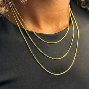May include: Three gold-toned curb chain necklaces of varying lengths, each featuring a 1.5mm link width. The necklaces are layered, displaying a consistent metallic yellow finish and a classic interlocking link design against a black fabric background.