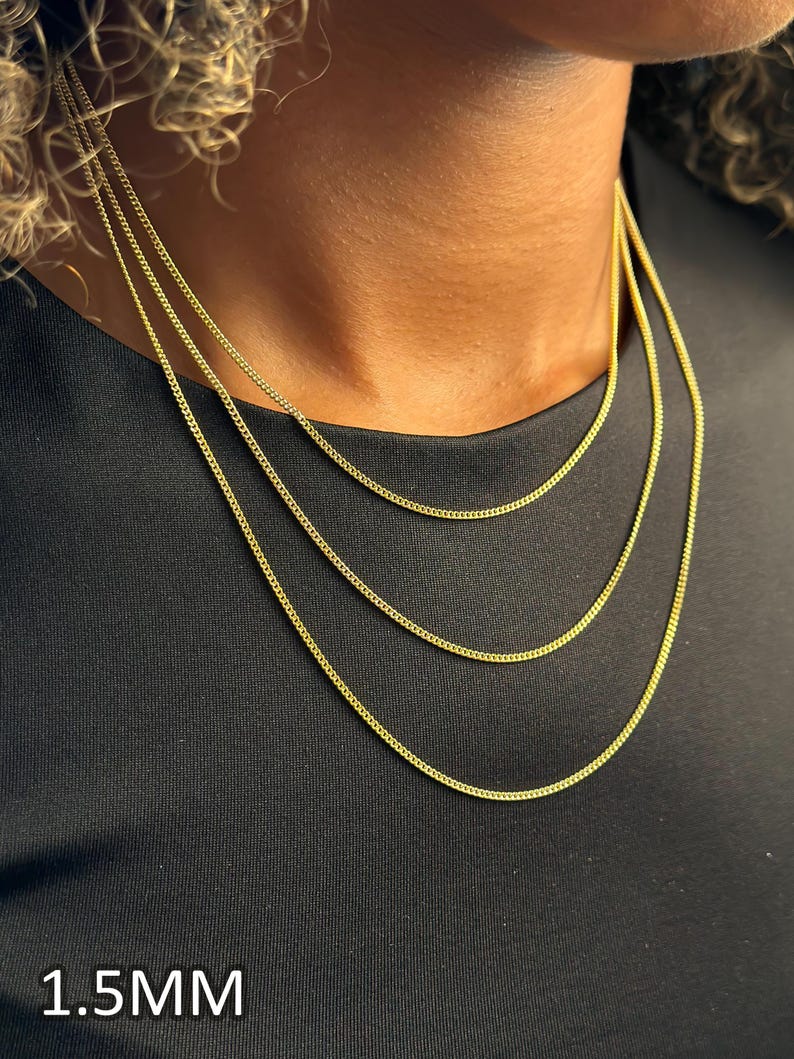 May include: Three gold-toned curb chain necklaces of varying lengths, each featuring a 1.5mm link width. The necklaces are layered, displaying a consistent metallic yellow finish and a classic interlocking link design against a black fabric background.
