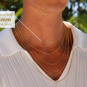 May include: A set of five layered gold Cuban chains worn around the neck. The text on the image indicates a Cuban chain with a 1.5mm width made of solid 14k gold. The chains are thin, metallic, and yellow in color, resting against a white shirt.