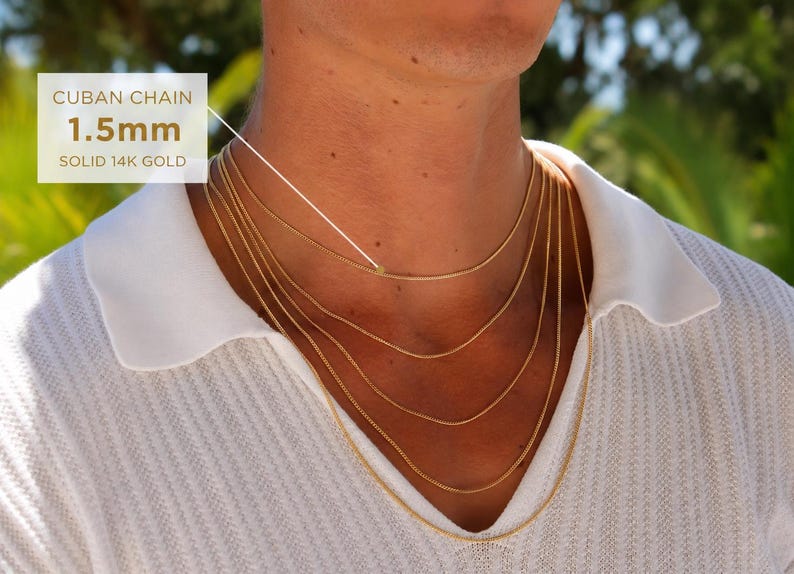 May include: A set of five layered gold Cuban chains worn around the neck. The text on the image indicates a Cuban chain with a 1.5mm width made of solid 14k gold. The chains are thin, metallic, and yellow in color, resting against a white shirt.