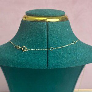 May include: A delicate gold necklace with a small clasp and chain is displayed on a green velvet jewelry bust. The necklace is shown against a soft pink background, highlighting its fine details and elegant design.