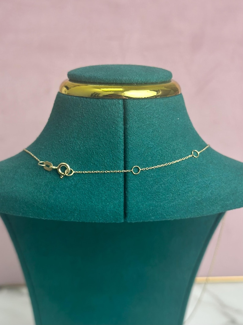 May include: A delicate gold necklace with a small clasp and chain is displayed on a green velvet jewelry bust. The necklace is shown against a soft pink background, highlighting its fine details and elegant design.