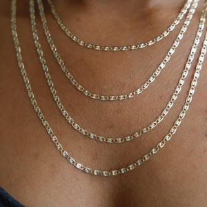 Puede incluir: Se muestran cuatro collares de cadena dorados de diferentes longitudes. Cada cadena presenta un dise&ntilde;o de eslabones ovalados repetidos. Los collares est&aacute;n superpuestos, creando un efecto en cascada. Un tatuaje de mariposa es visible en la piel.