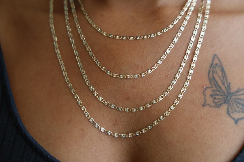 Puede incluir: Se muestran cuatro collares de cadena dorados de diferentes longitudes. Cada cadena presenta un dise&ntilde;o de eslabones ovalados repetidos. Los collares est&aacute;n superpuestos, creando un efecto en cascada. Un tatuaje de mariposa es visible en la piel.