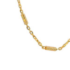 Puede incluir: Un collar de cadena dorada que presenta una serie de eslabones ovalados conectados por cuentas cil&iacute;ndricas de oro texturizado. El metal tiene un acabado brillante y reflectante, alternando entre eslabones lisos y cuentas con ranuras.