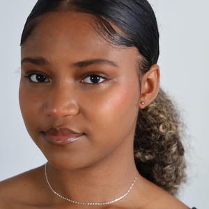 Puede incluir: Una mujer que lleva un delicado collar de plata. El collar es una cadena fina con eslabones peque&ntilde;os y espaciados uniformemente. Tambi&eacute;n lleva un peque&ntilde;o pendiente de oro. La mujer tiene el pelo oscuro recogido y lleva una camiseta negra.