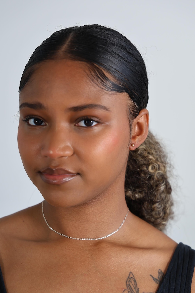 Puede incluir: Una mujer que lleva un delicado collar de plata. El collar es una cadena fina con eslabones peque&ntilde;os y espaciados uniformemente. Tambi&eacute;n lleva un peque&ntilde;o pendiente de oro. La mujer tiene el pelo oscuro recogido y lleva una camiseta negra.