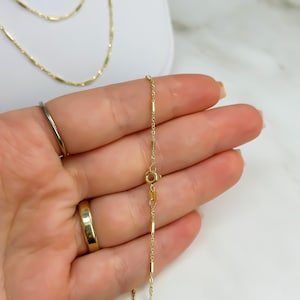 Puede incluir: Un delicado collar de cadena de oro con peque&ntilde;os eslabones de barra cil&iacute;ndricos espaciados uniformemente. La cadena incluye un cierre de resorte circular y una peque&ntilde;a etiqueta rectangular cerca del cierre, con un acabado met&aacute;lico pulido.