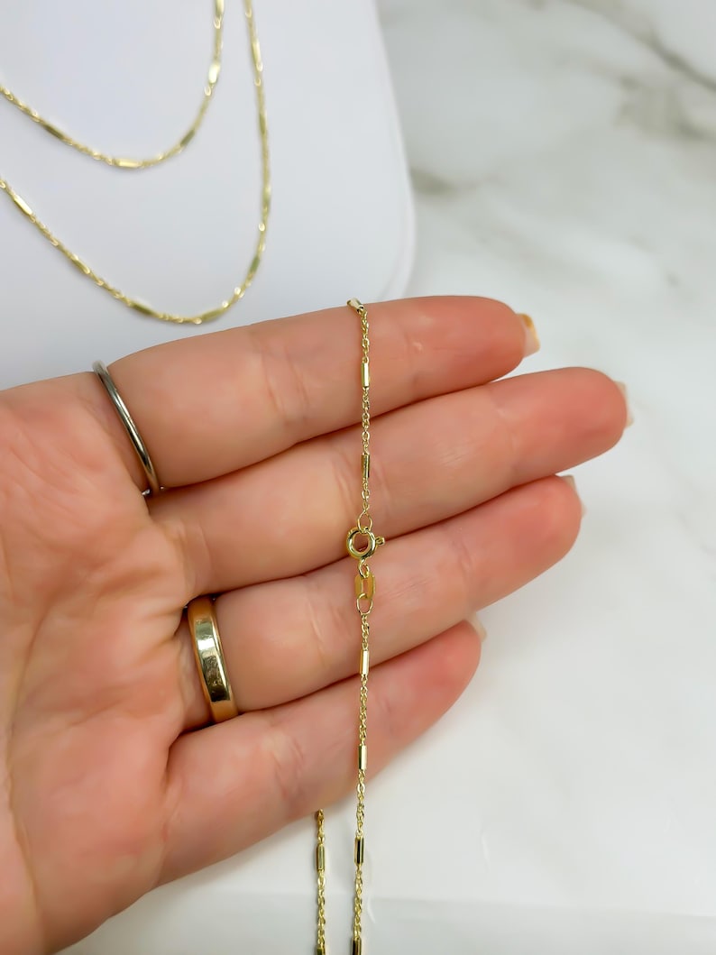 Puede incluir: Un delicado collar de cadena de oro con peque&ntilde;os eslabones de barra cil&iacute;ndricos espaciados uniformemente. La cadena incluye un cierre de resorte circular y una peque&ntilde;a etiqueta rectangular cerca del cierre, con un acabado met&aacute;lico pulido.