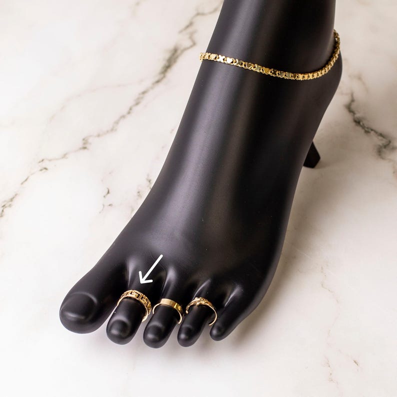 May include: Gold toe rings on a black mannequin foot. The rings are worn on the big toe, second toe, and third toe. The rings are simple and delicate.