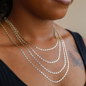 Puede incluir: Un conjunto de collares en capas con cuatro delicadas cadenas planas doradas. Las brillantes cadenas descansan sobre el pecho de una persona que lleva una camiseta negra. El fondo es de color neutro.