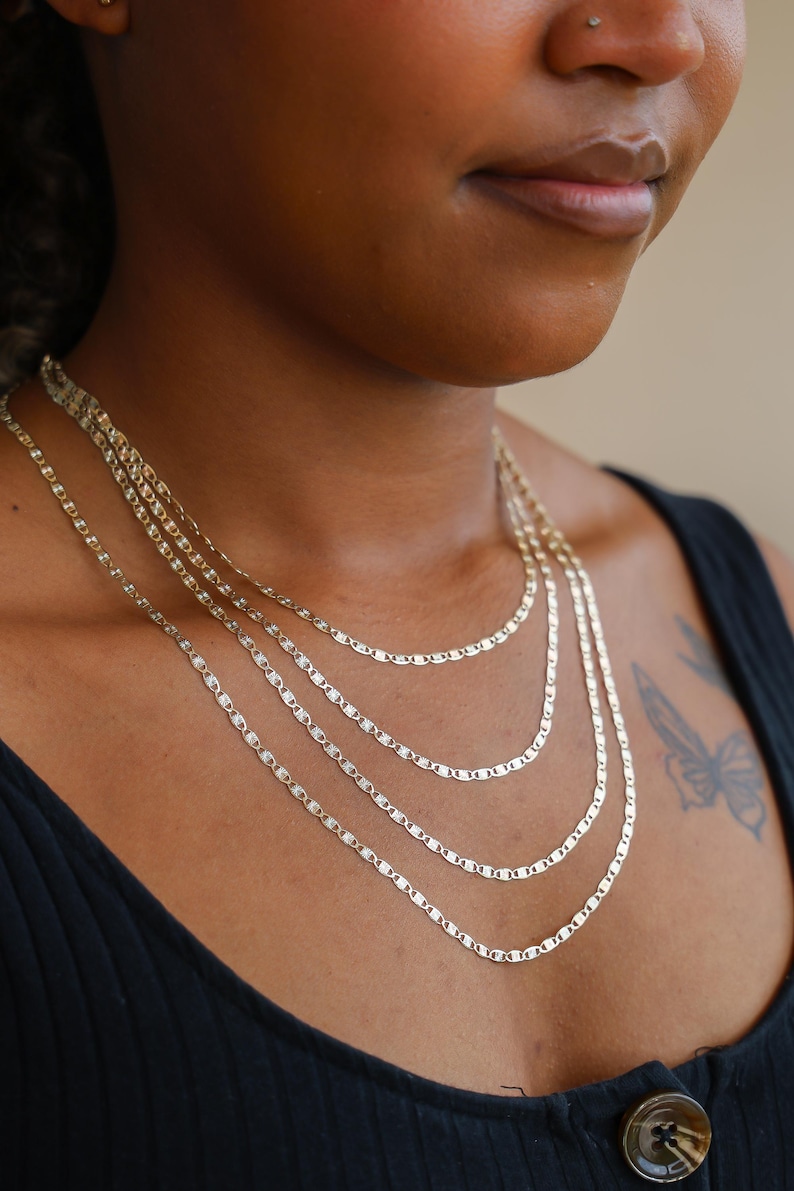 Puede incluir: Un conjunto de collares en capas con cuatro delicadas cadenas planas doradas. Las brillantes cadenas descansan sobre el pecho de una persona que lleva una camiseta negra. El fondo es de color neutro.