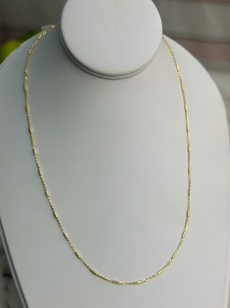 Puede incluir: Un delicado collar de cadena de oro que presenta una serie de peque&ntilde;os eslabones cil&iacute;ndricos espaciados uniformemente, conectados por finos eslabones circulares de alambre. La cadena tiene un acabado met&aacute;lico brillante y se muestra en un bucle.