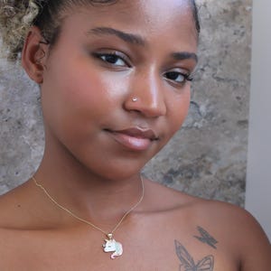 May include: A gold necklace with a unicorn pendant. The pendant features a white unicorn with a pink mane and tail, and a blue horn. The person in the image has a butterfly tattoo on their shoulder.
