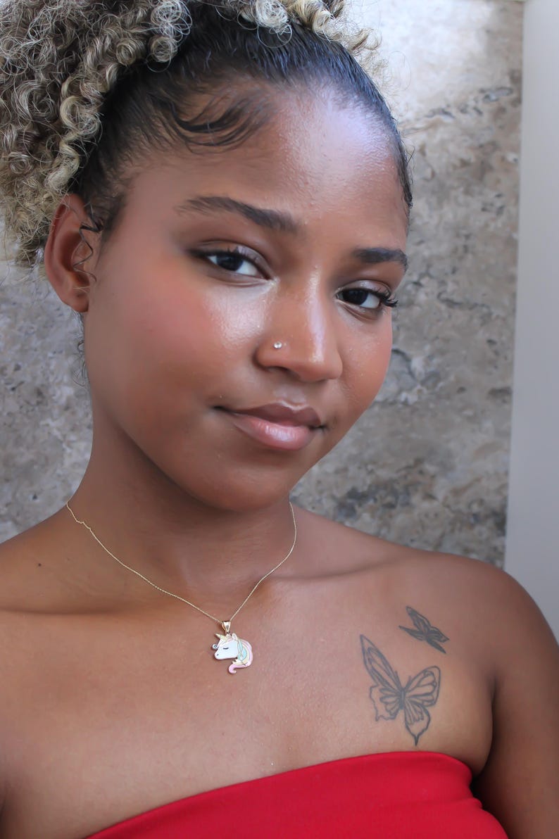 May include: A gold necklace with a unicorn pendant. The pendant features a white unicorn with a pink mane and tail, and a blue horn. The person in the image has a butterfly tattoo on their shoulder.
