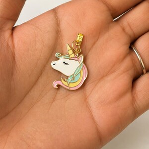 May include: A gold-colored unicorn charm with a white face, a yellow horn, and pink, blue, and yellow mane. The charm is held in a hand, and the background is a light color.