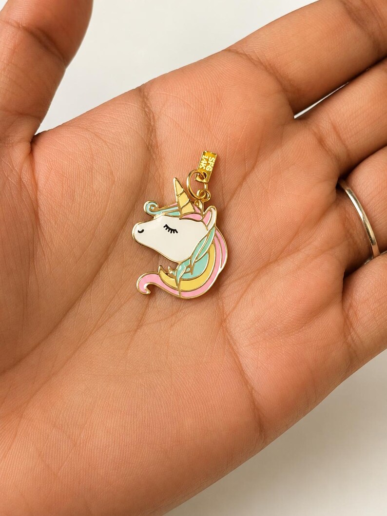 May include: A gold-colored unicorn charm with a white face, a yellow horn, and pink, blue, and yellow mane. The charm is held in a hand, and the background is a light color.