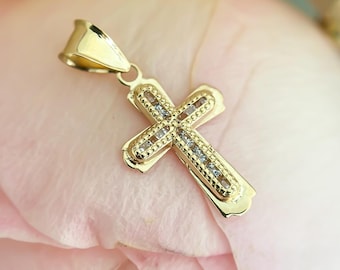 May include: A gold cross pendant featuring a central channel set with small clear stones. The cross has a polished gold border with a beaded texture along the inner edges and includes a tapered gold bail at the top for attachment to a chain.